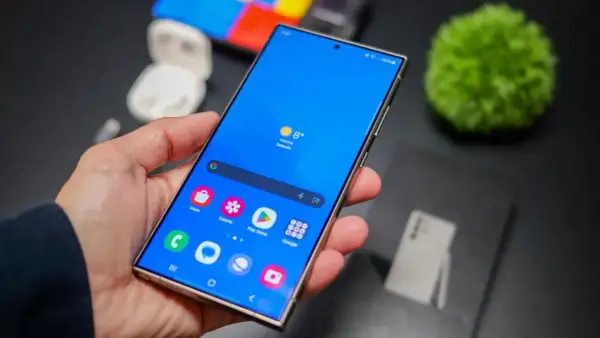 Mano sujetando un smartphone con pantalla azul vibrante, mostrando iconos de aplicaciones y el tiempo. La interfaz, **que es One UI**, se ve fluida y organizada.